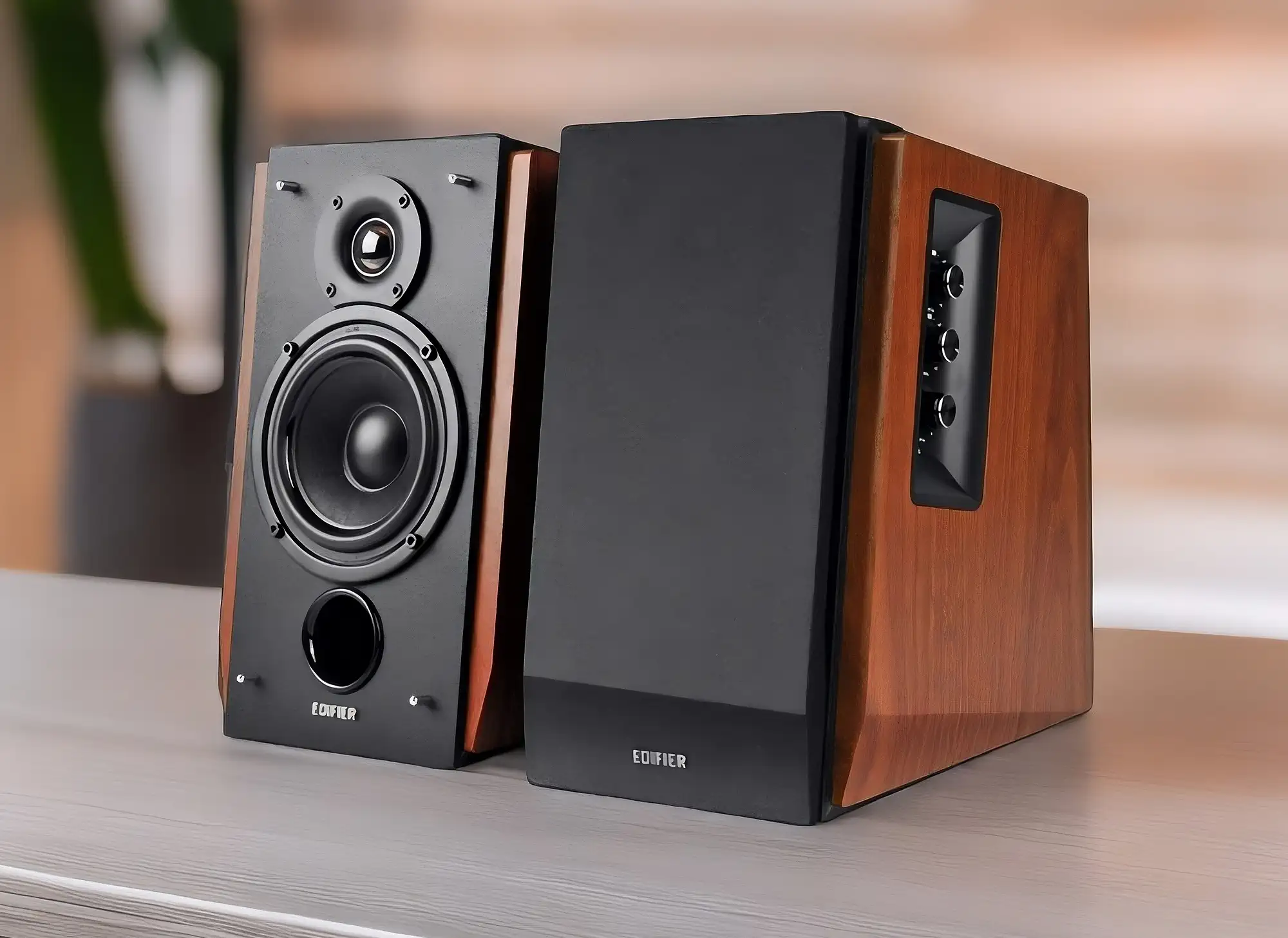 Колонки Edifier R1700BT на светлом столе. Модели с наклонным корпусом и отделкой боковых панелей под дерево. У левой колонки снят гриль, открывая вид на динамики; правая – под защитной сеткой. На боковой грани активной колонки расположены три регулятора.