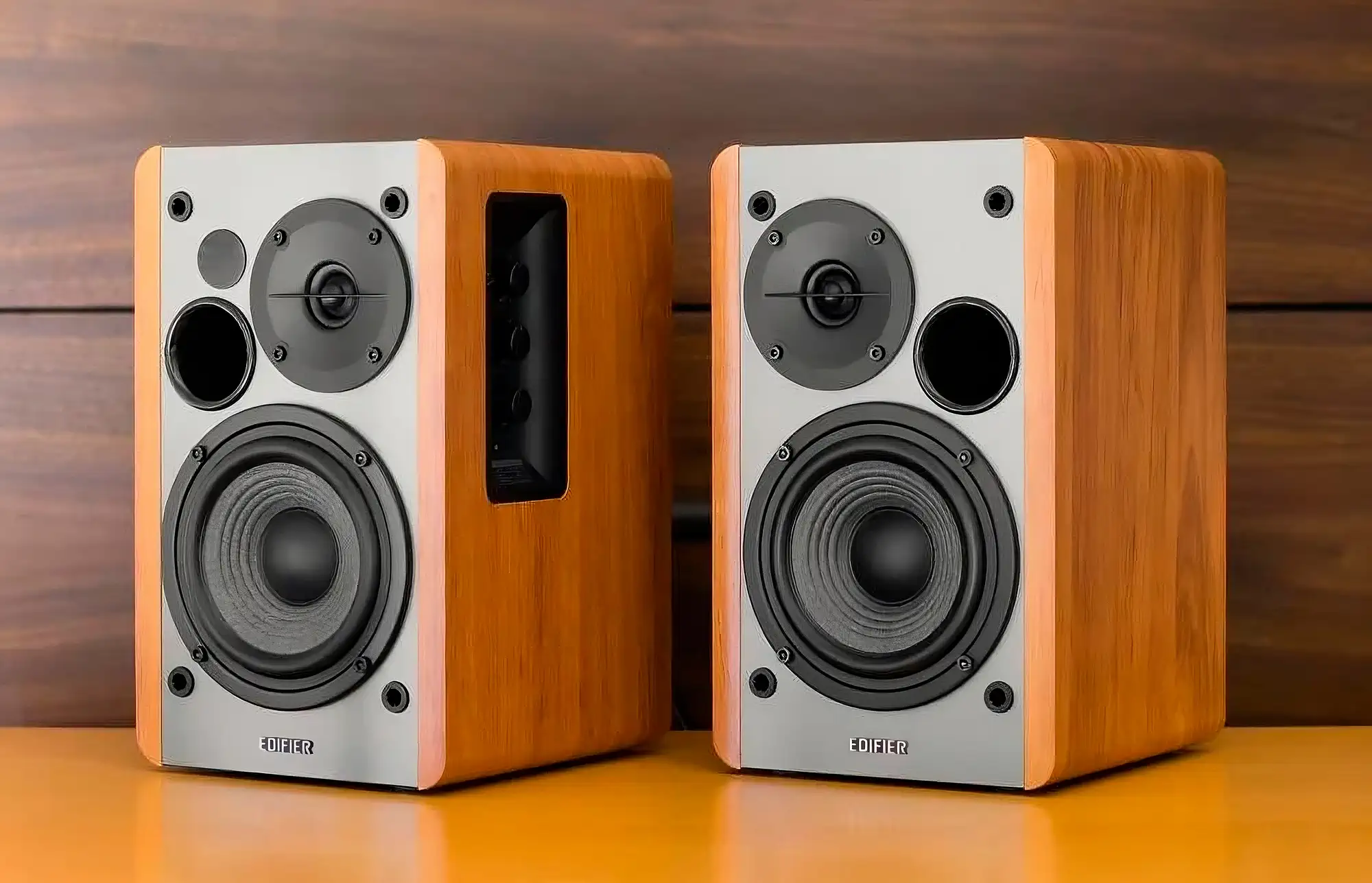 Колонки Edifier R1280DBs на деревянном столе. Модели с серыми передними панелями и светлыми боковыми стенками под дерево; грили сняты, открывая вид на динамики и фронтальные порты фазоинвертора. На боковой грани активной колонки расположена ниша с регуляторами. Фронтальный вид.