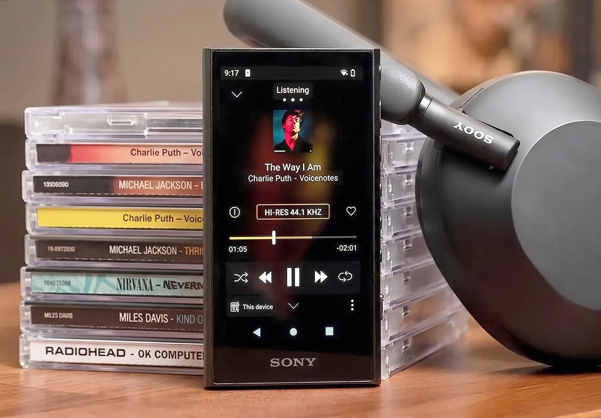 Плеер Sony NW-A306. Устройство с включенным экраном стоит в центре, слева – стопка CD, справа наушники Sony. Лайфстайл-фото на деревянном столе.