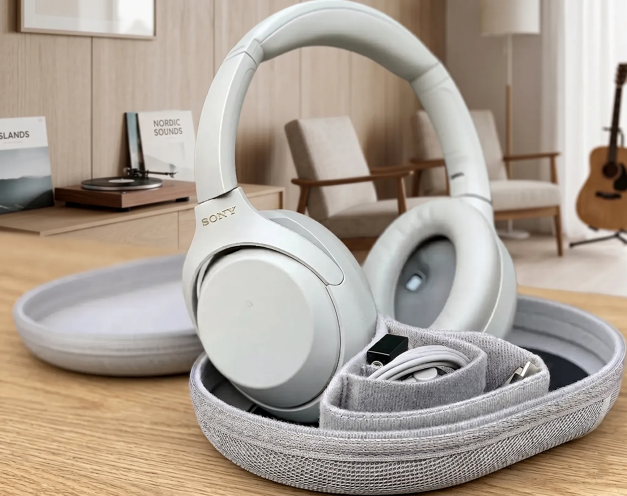 Sony WH-1000XM4 в открытом кейсе с кабелем на столе, фон – комната с гитарой и креслами, светлый план.