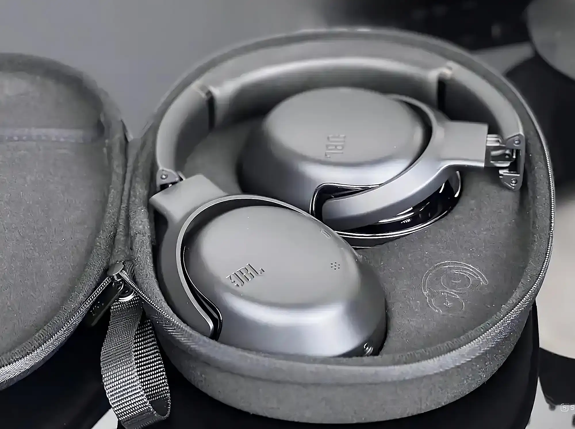 JBL Tour One M2 в открытом сером футляре: сложенные чашки, вид сверху под углом, матовый корпус.