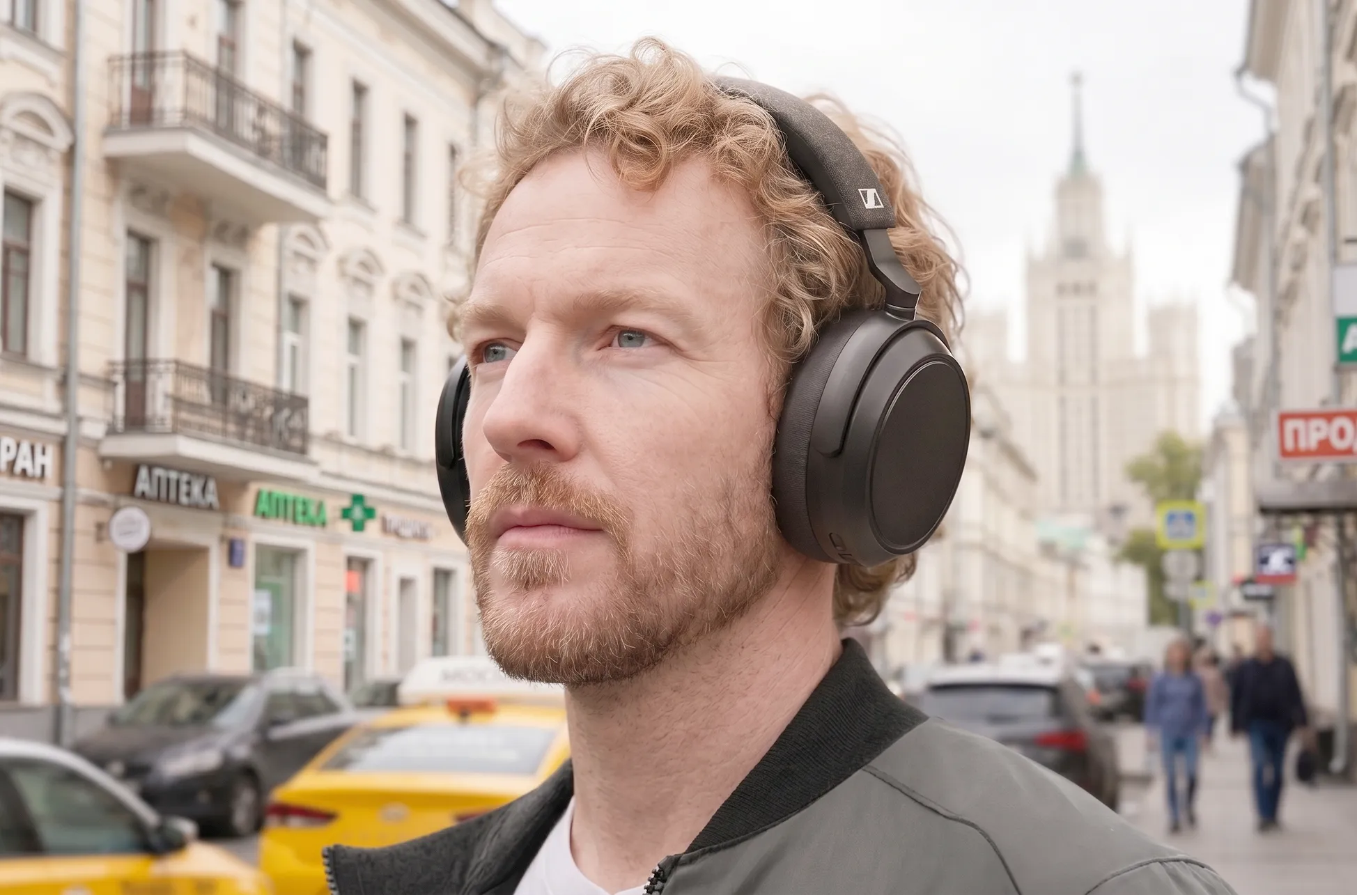 Павел Окопень в Sennheiser MOMENTUM 4: портрет на улице с вывесками, черные чашки и тканевое оголовье.