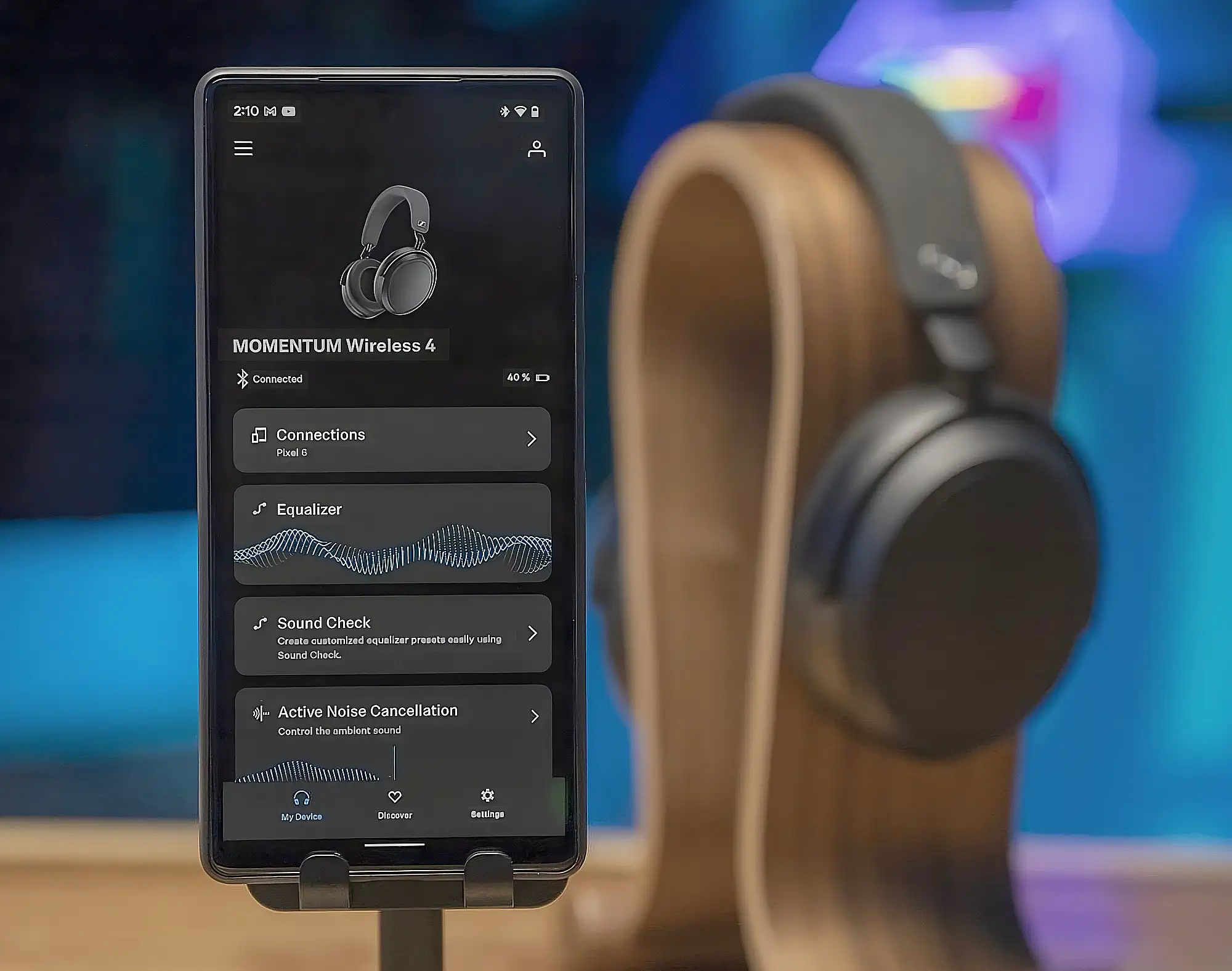 Sennheiser Momentum 4 и Smart Control: смартфон с меню настроек на переднем плане, наушники на стойке сзади.