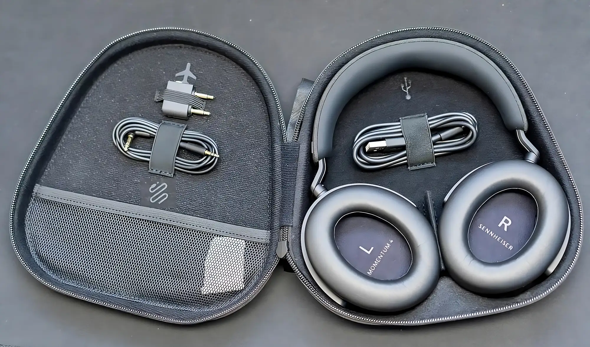 Sennheiser Momentum 4 в открытом кейсе: адаптер и кабель слева, чашки L и R справа, вид сверху, темный цвет.