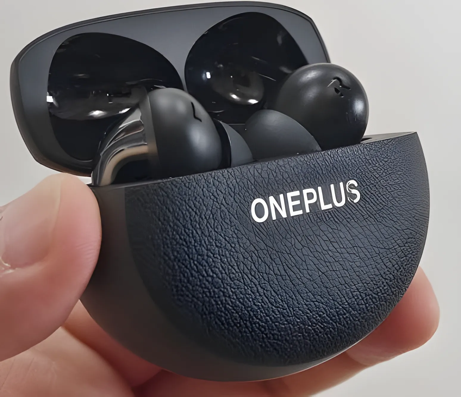 OnePlus Buds Pro 3: открытый кейс с наушниками в руке, черный текстурный футляр с серебристым логотипом ONEPLUS.
