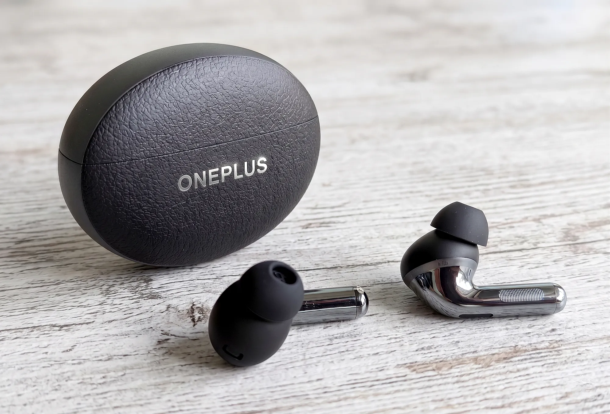OnePlus Buds Pro 3 – TWS–наушники и зарядный кейс на светлом деревянном столе. Овальный футляр с текстурой под кожу и серебристым логотипом, наушники с комбинированным матово–глянцевым корпусом. Крупный план, вид спереди.