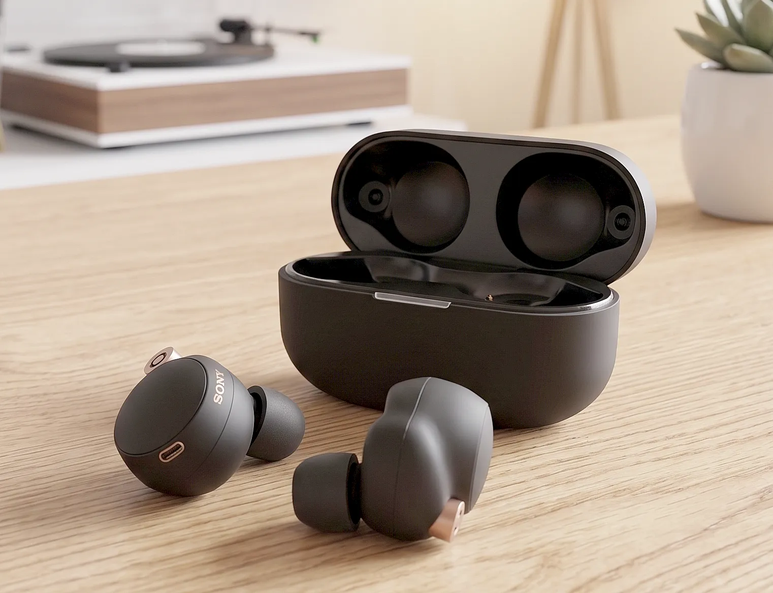 Sony WF-1000XM4: черные наушники и открытый кейс на светлом столе, матовый корпус с медными акцентами, фон – винил и суккулент.