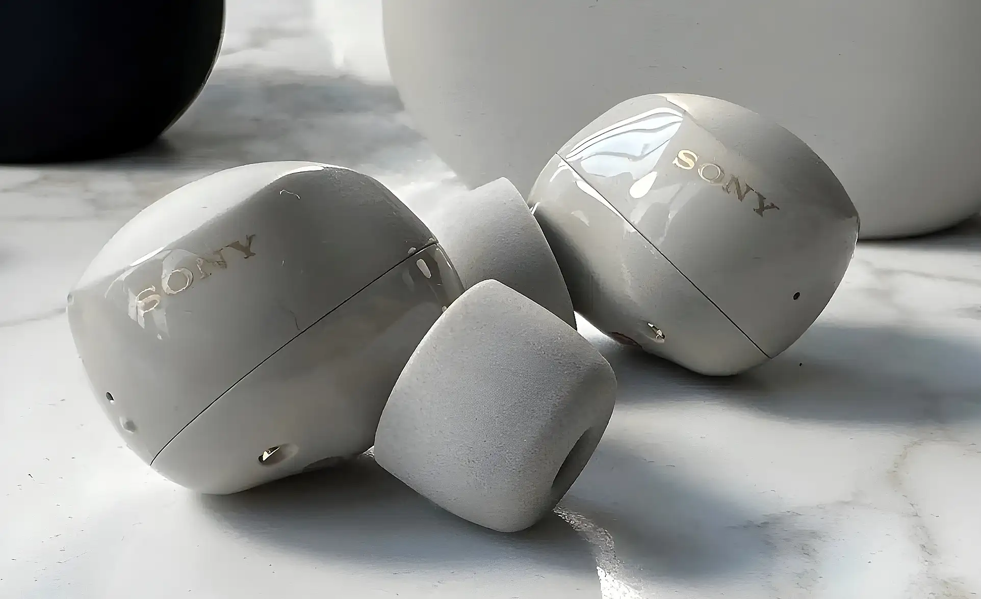 Sony WF-1000XM5: светло-серые наушники на мраморе, глянцевый корпус с золотистым логотипом SONY и пенными амбушюрами, макро.