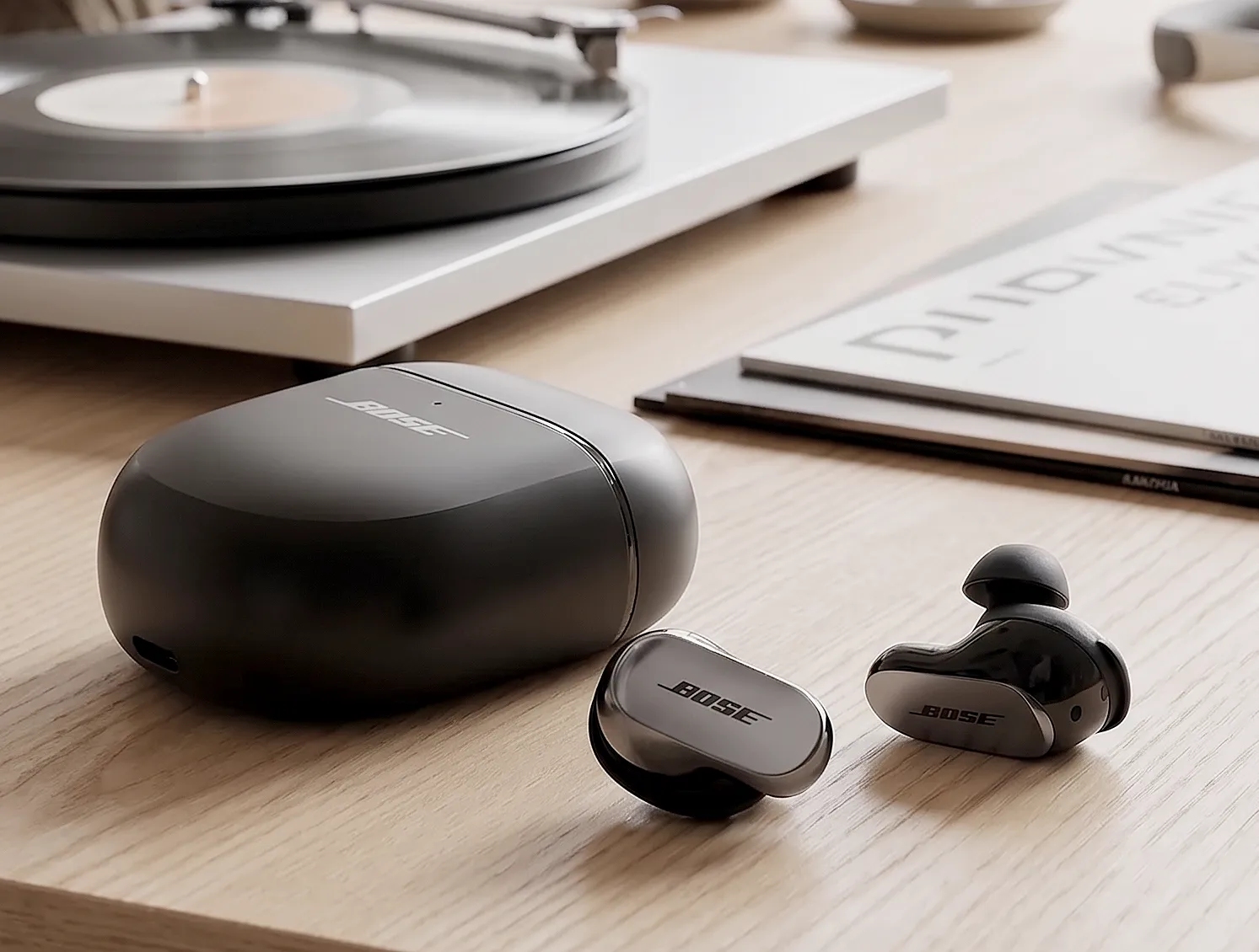 Bose QC Ultra Earbuds: черные наушники и закрытый кейс на светлом столе, глянцевая панель BOSE и матовый футляр.