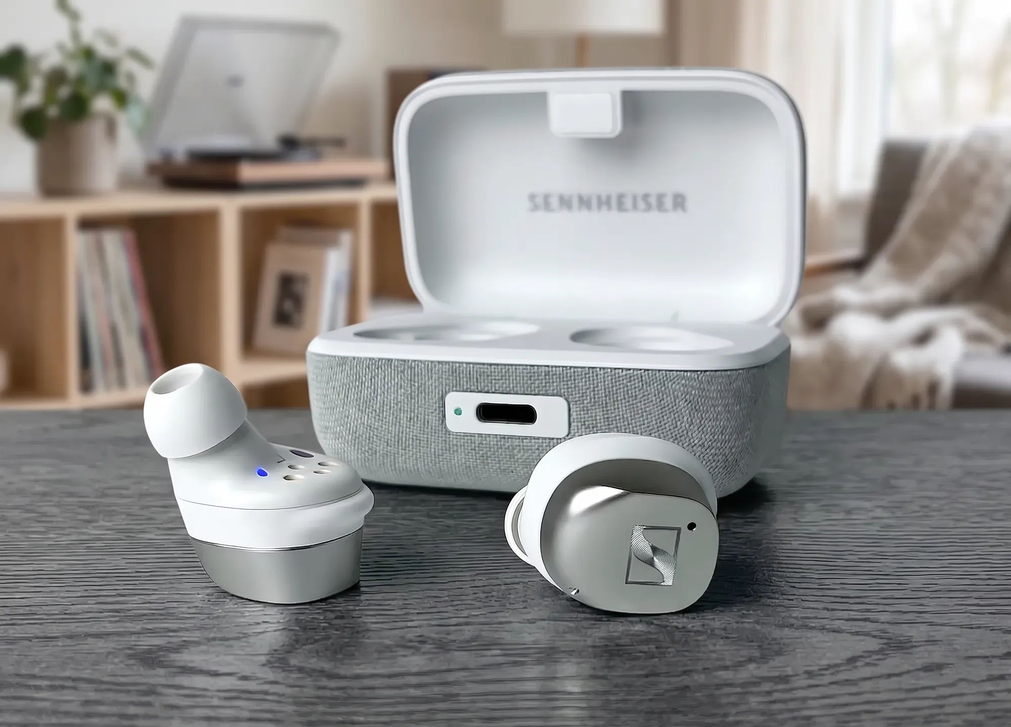Sennheiser Momentum True Wireless 4 – TWS–наушники и открытый зарядный кейс на темном деревянном столе. Футляр имеет фирменную отделку серым текстилем, на передней панели расположены порт USB–C и светодиодный индикатор. Внутриканальные наушники выполнены в комбинированном дизайне с серебристыми металлическими фейсплейтами и логотипом бренда.