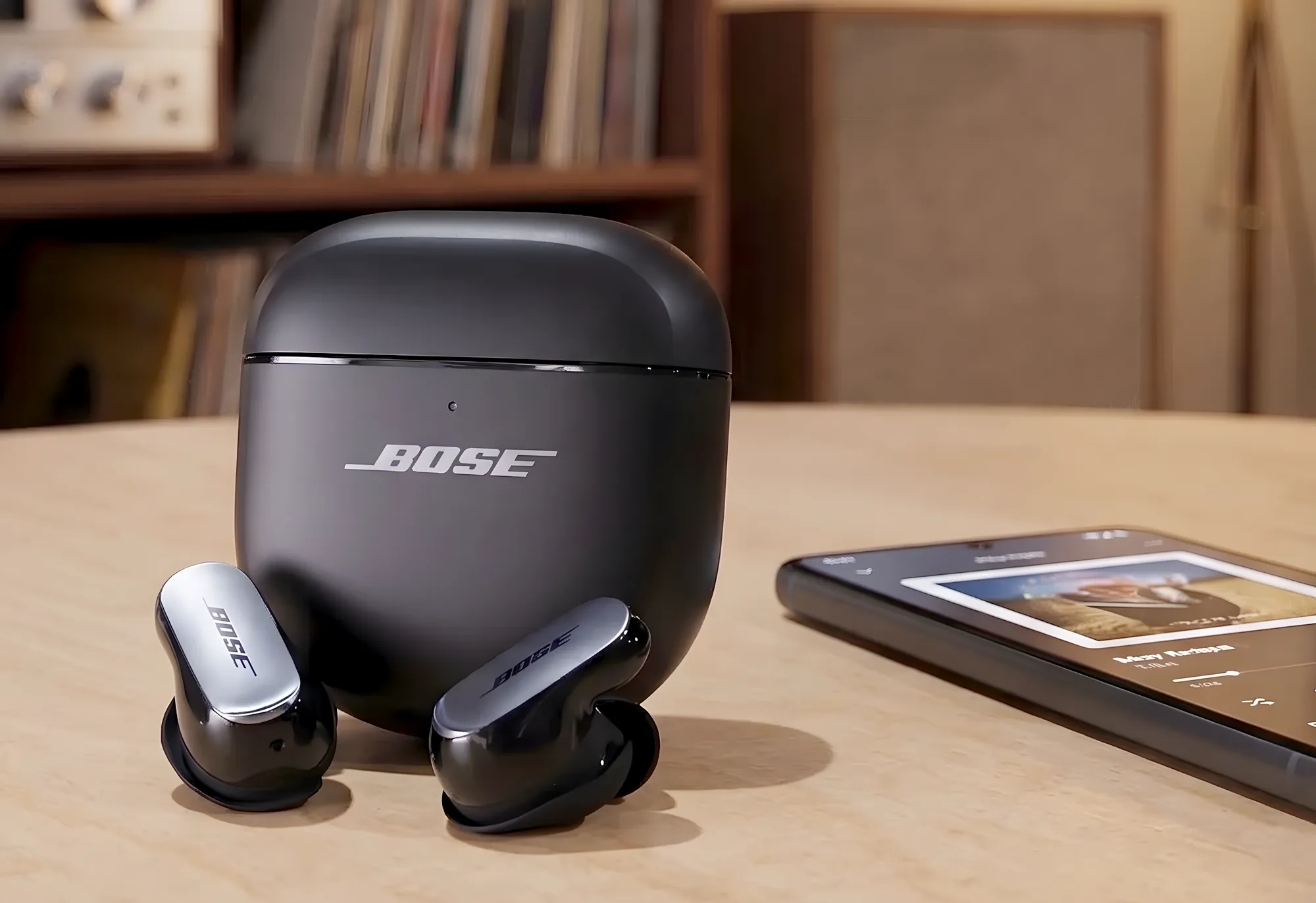 Bose QC Ultra Earbuds – черные TWS–наушники и закрытый зарядный кейс на светлом деревянном столе. Матовый футляр с серебристым логотипом «BOSE» стоит вертикально, перед ним лежат наушники с глянцевым корпусом и металлизированными фейсплейтами.
