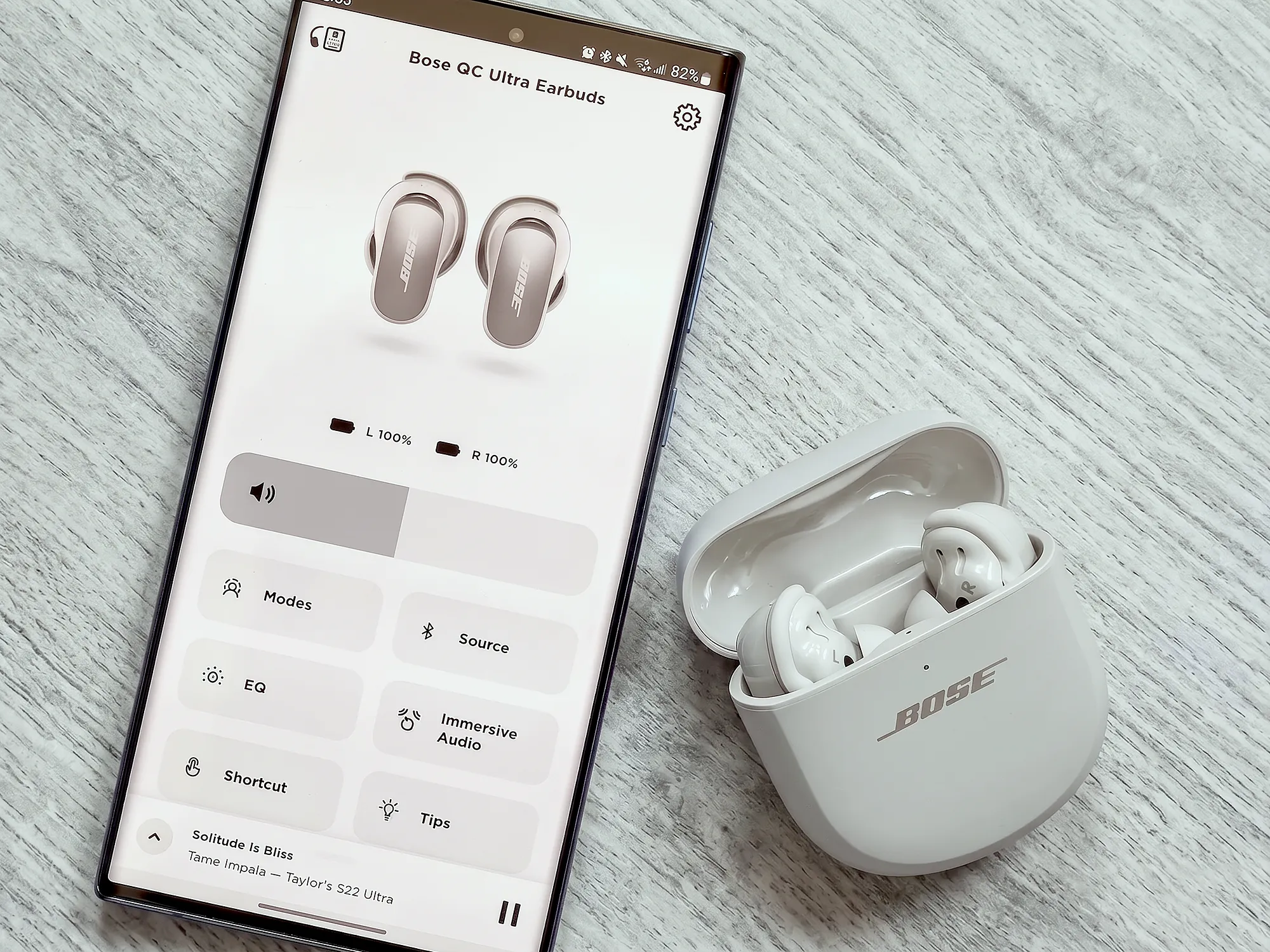Bose QC Ultra Earbuds: смартфон с приложением, открытый белый кейс и наушники на светлом столе, интерфейс Modes, EQ и Immersive Audio.