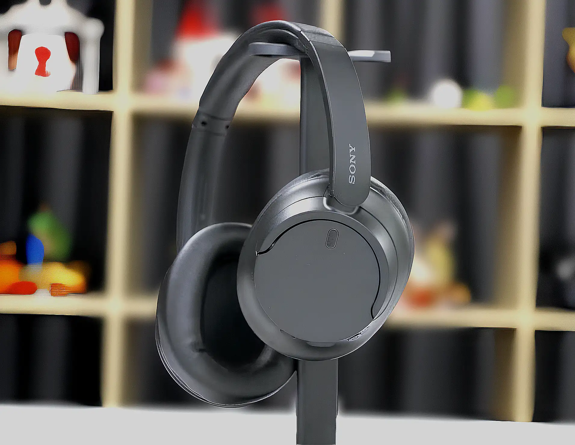 Полноразмерные наушники Sony WH–CH720N черного цвета висят на стойке. Овальная чашка с логотипом бренда крупным планом на размытом фоне.