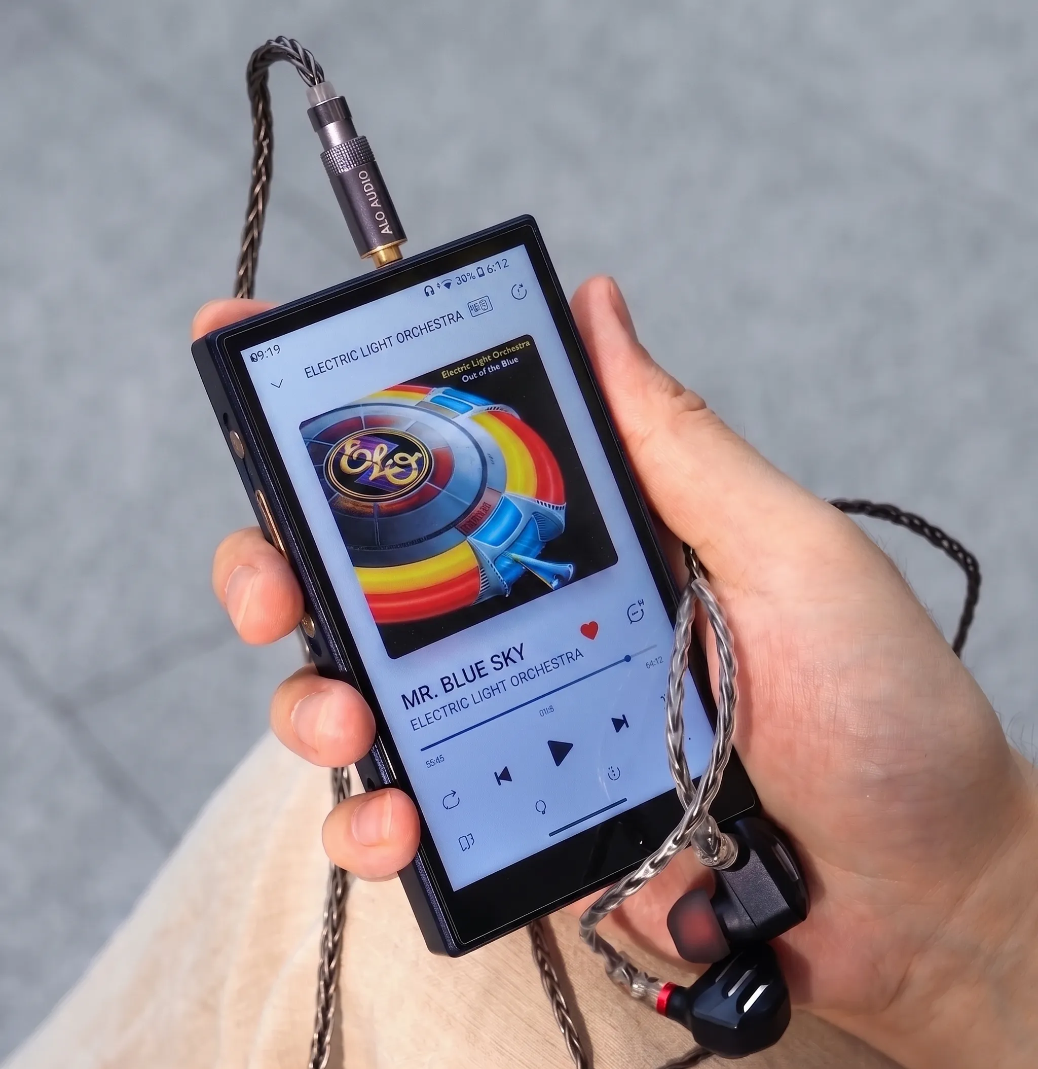 Аудиоплеер FiiO M21 в руке: экран с кассетным интерфейсом, подключенный кабель сверху и наушники.