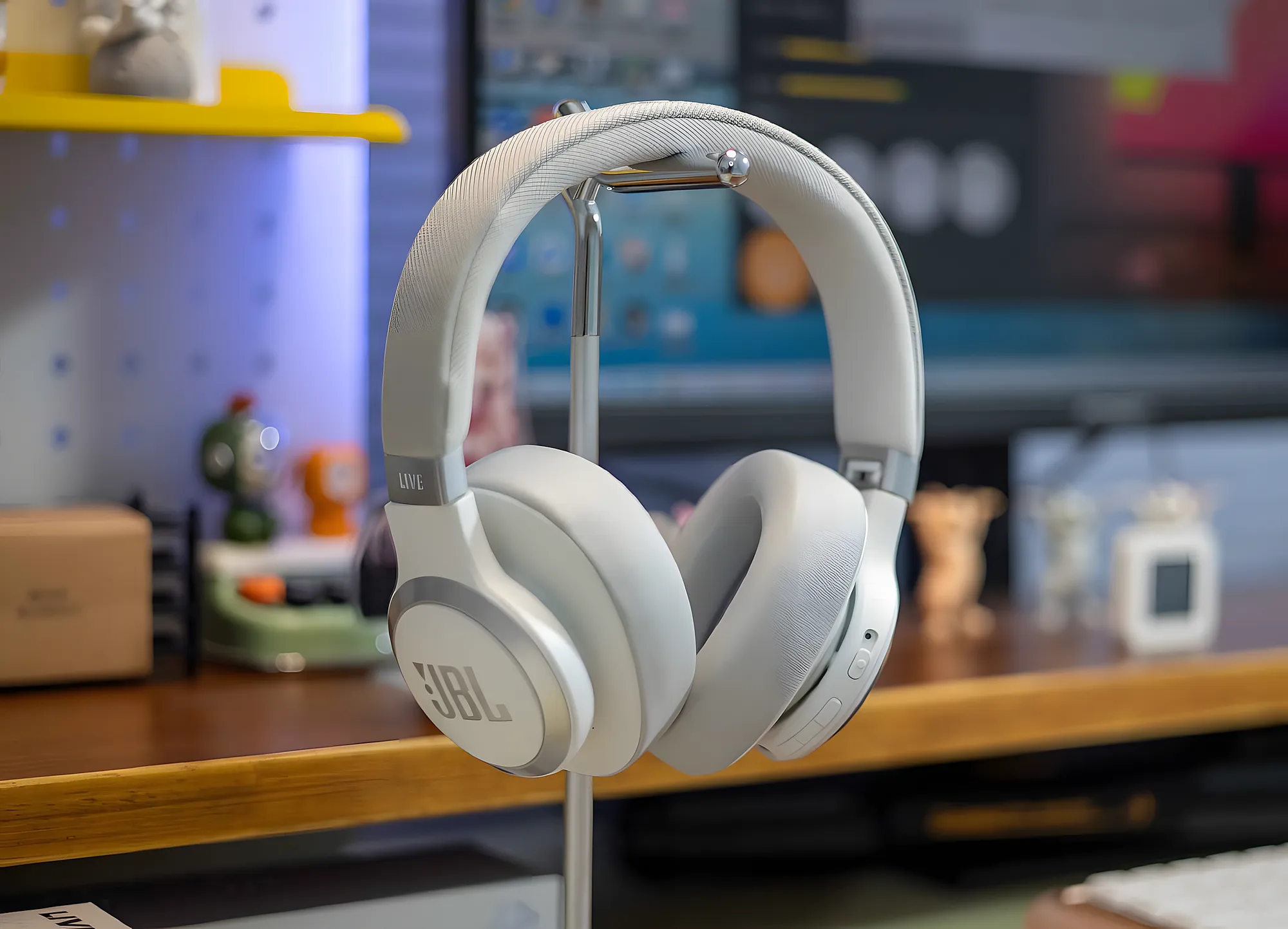 Беспроводные наушники JBL Live 770NC в светлом цвете висят на металлической стойке перед рабочим столом, фон с монитором и аксессуарами слегка размыт.