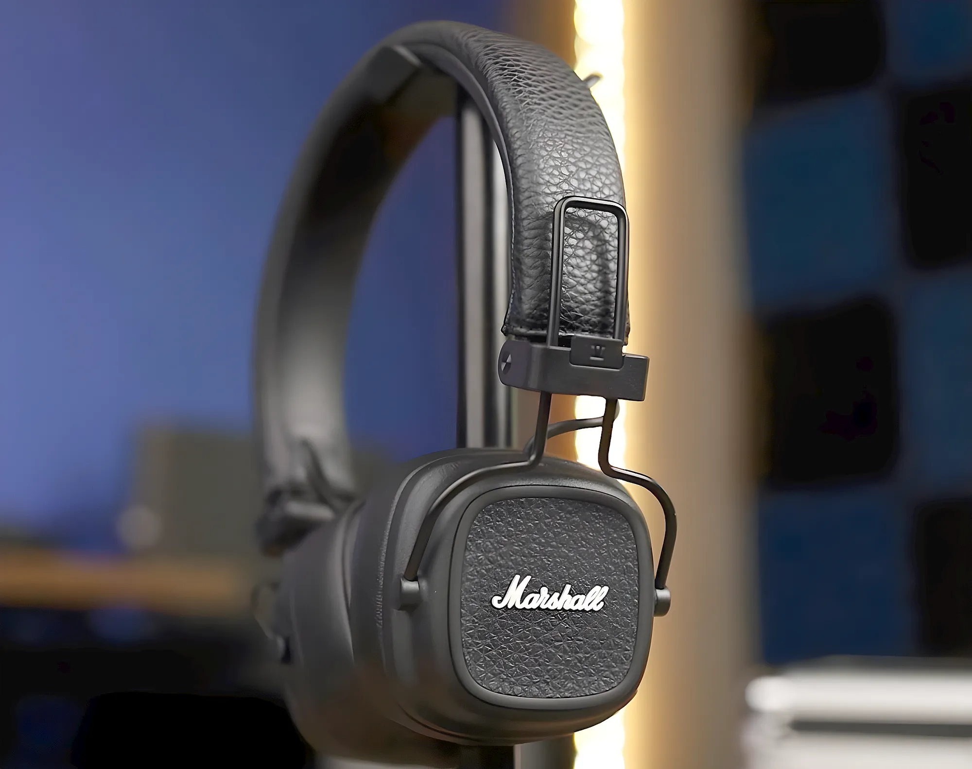 Беспроводные наушники Marshall Major V висят на настольной стойке, левая чашка с логотипом развернута к камере, фон слегка размытый с синими панелями.