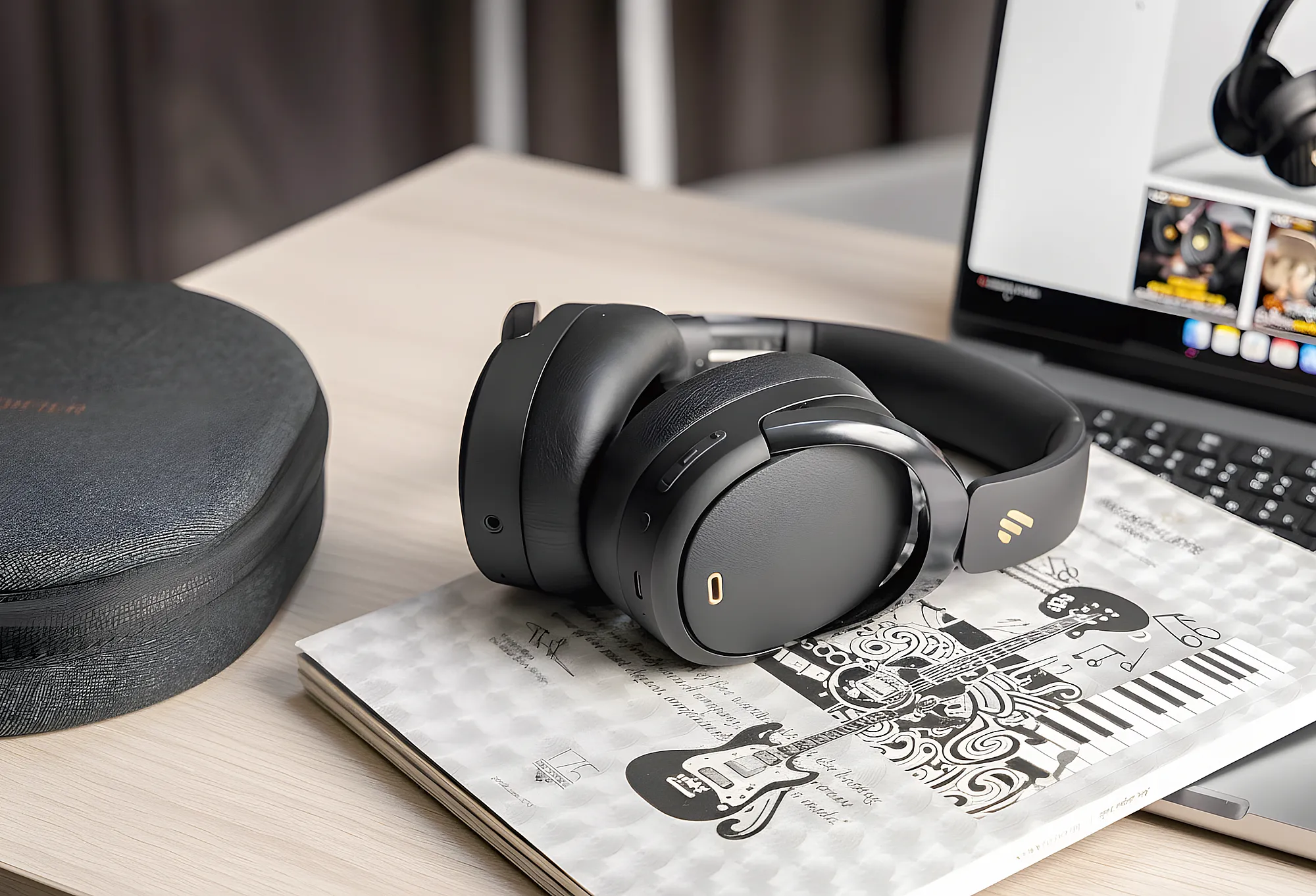 Беспроводные наушники Edifier WH950NB лежат на иллюстрированном блокноте на столе, слева чехол, справа частично виден ноутбук с открытым экраном