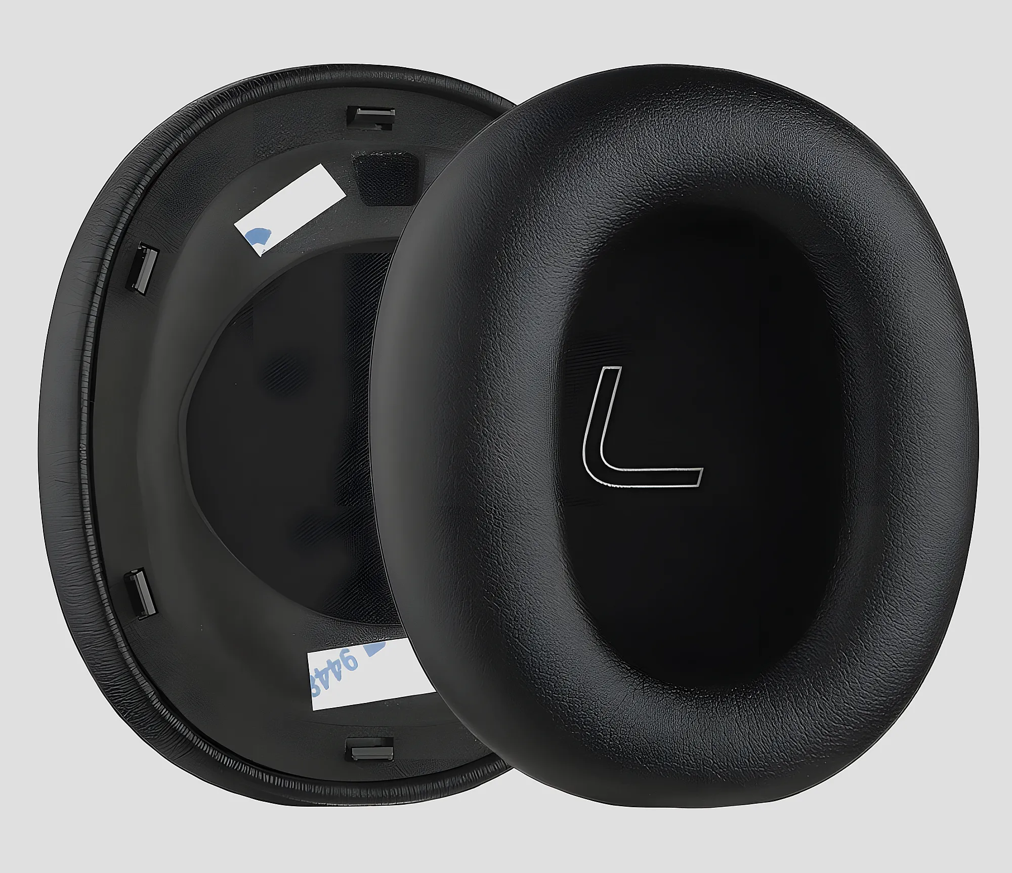 Пара овальных сменных амбушюр из черной эко-кожи с эффектом памяти, одна перевернута для демонстрации креплений и клеевой площадки, фон однотонный серый.