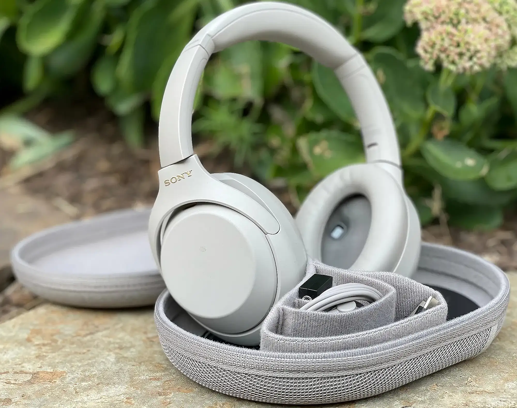 Ответ Gemini  Наушники Sony WH-1000XM4 бежевого цвета в открытом кейсе на камне. Видны мягкие амбушюры, золотистый логотип, кабель и адаптер в чехле.